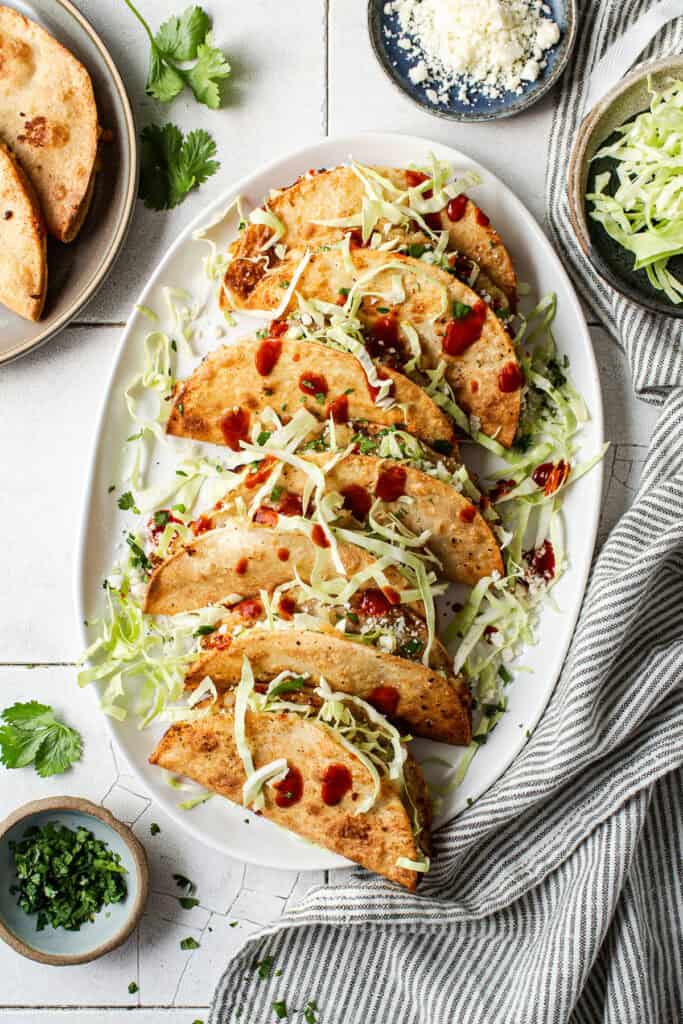 A serving plate of tacos de papa, aka crispy potato tacos, all stuffed with shredded cabbage, cotija, and drizzles of hot sauce.