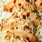 Close-up of a crispy potato taco with cabbage, cotija, and hot sauce on a plate.