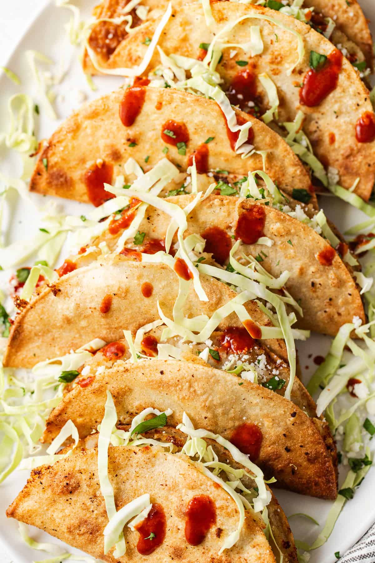 Close-up of a crispy potato taco with cabbage, cotija, and hot sauce on a plate.